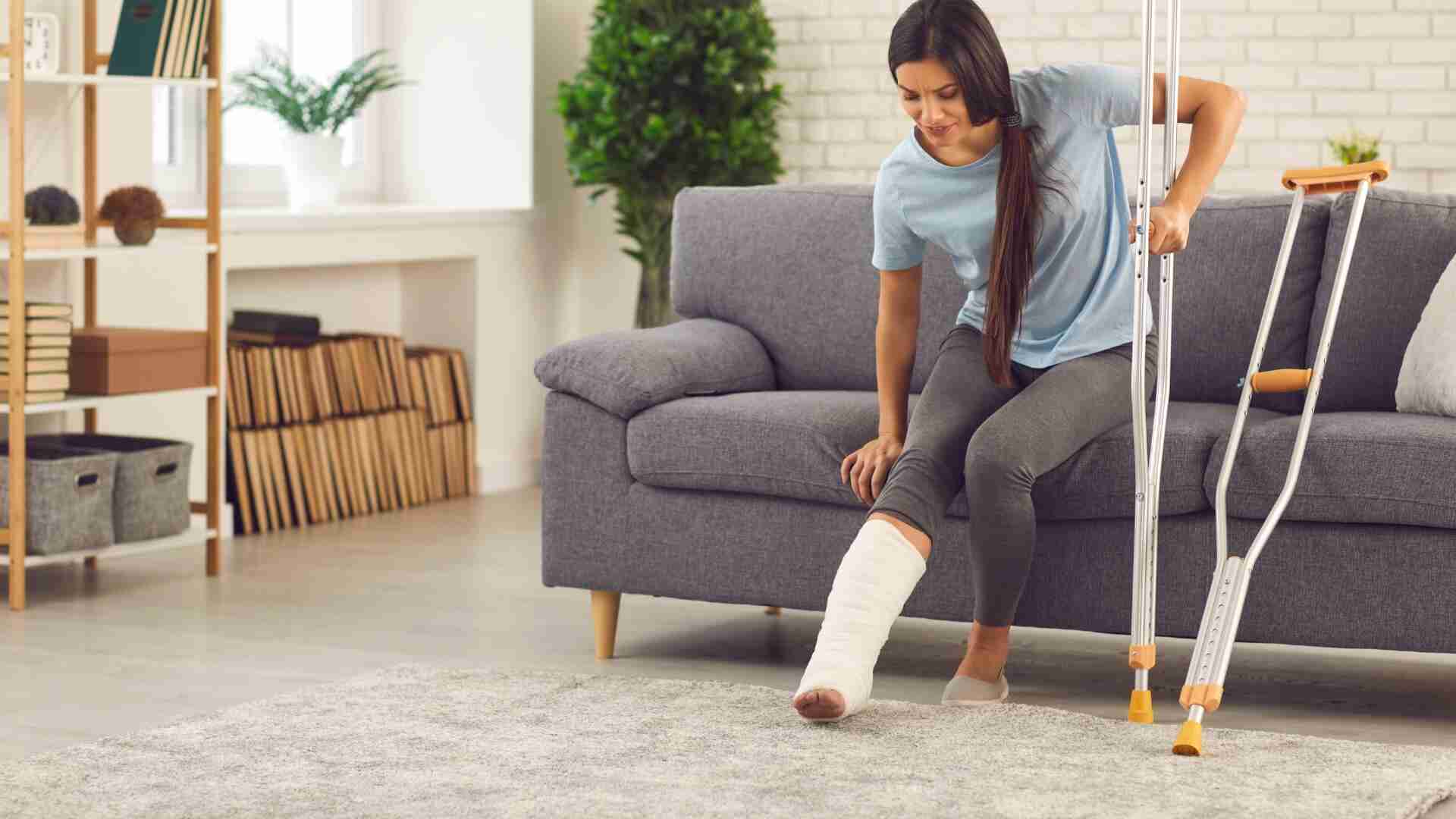 Frau sitzt auf einem Sofa und betrachtet ihren Gipsverband am Bein, während sie Krücken benutzt.